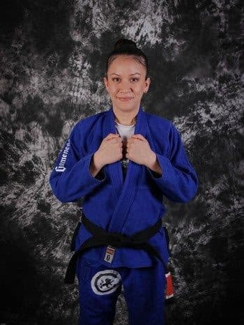 Studio portrait of a smiling woman in a blue gi wearing her Brazilian Jiu Jitsu black belt.