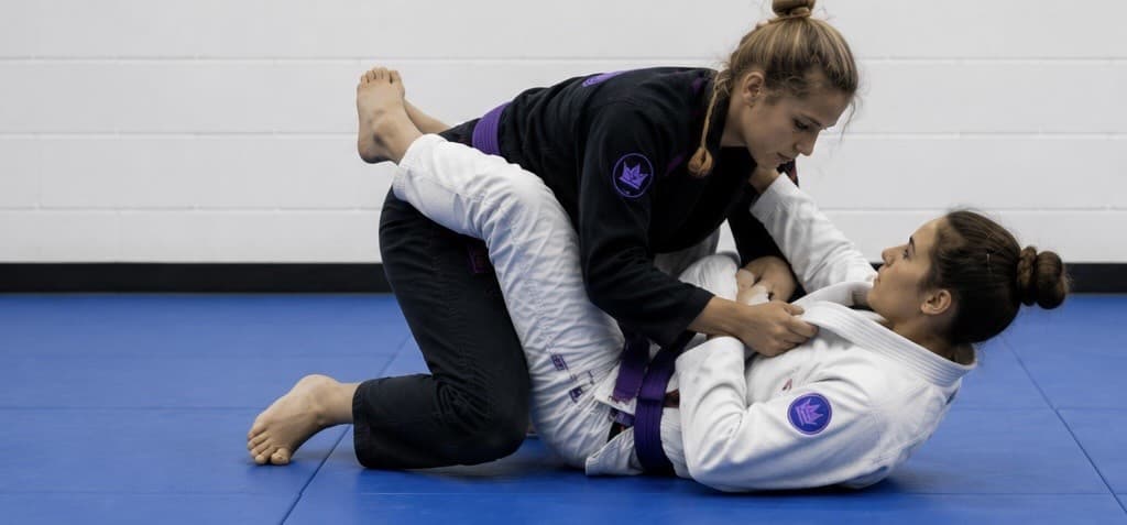 Two women training Brazilian Jiu Jitsu, a purple belt in a black gi attacking from the top while her partner in a white gi defends guard off her back.
