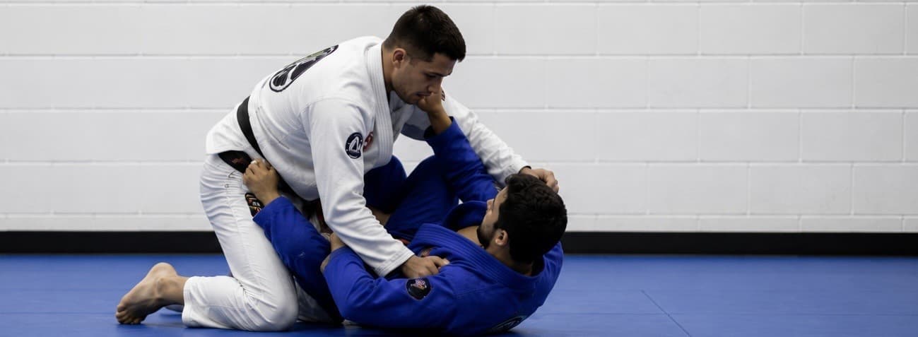 Two men sparring in an adult Brazilian Jiu Jitsu class, a black belt in a white gi working to pass the open guard of his training partner in a blue gi.